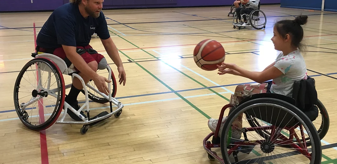 young lady in a wheelchair tossing a basketball to a gentleman to her left while on an indoor basketball court