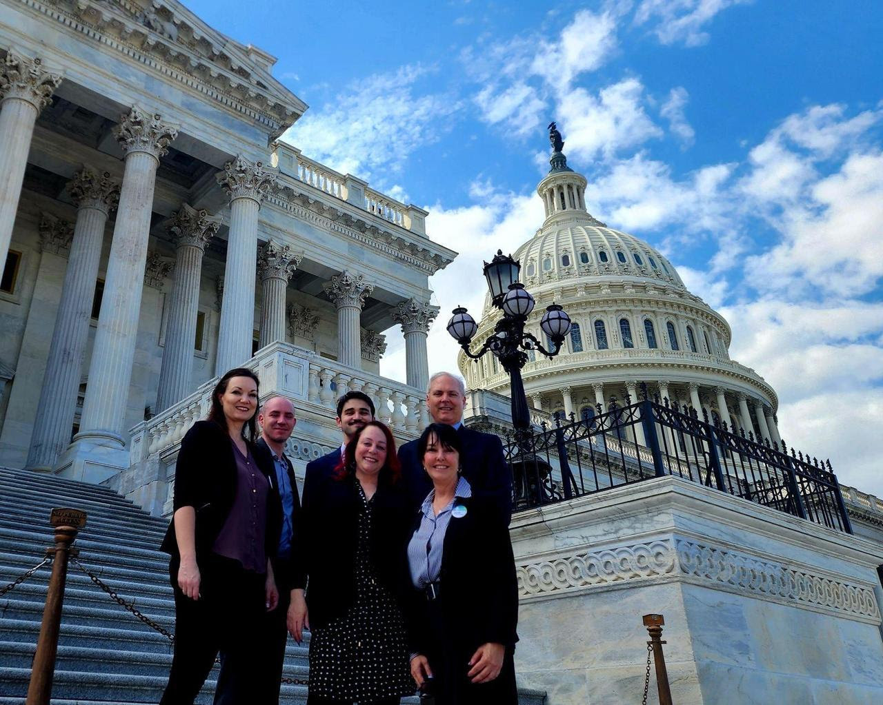 A group of disability advocates stand on the steps of Capitol Hill on under a blue sky on a sunny day