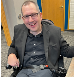 A man using a wheelchair wearing glasses and a sports coat smiles at the camera