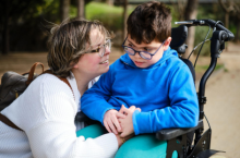 Woman with short brown hair and glasses talking to a child with glasses who is sitting in a wheelchair